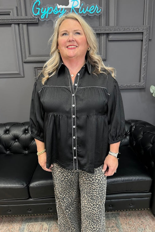 Woman wearing a black blouse and patterned pants standing in front of a black leather couch with 'Gypsy River' sign.
