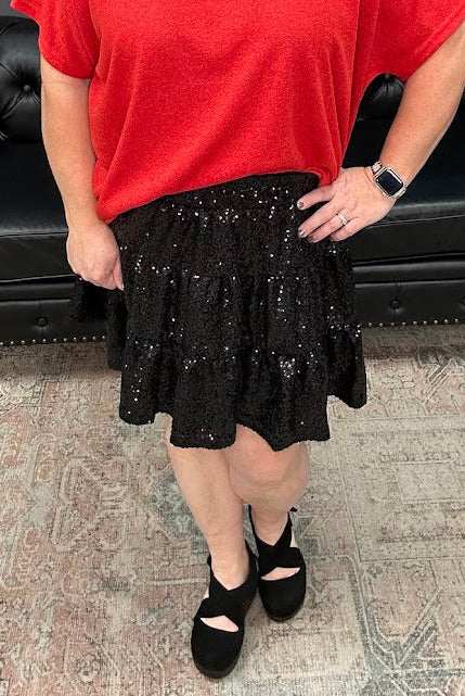 Woman wearing a red top and black skirt standing in a room with a black leather couch and decorative items.