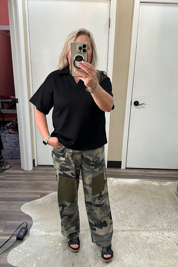 Person wearing a black polo shirt and camouflage pants taking a mirror selfie.