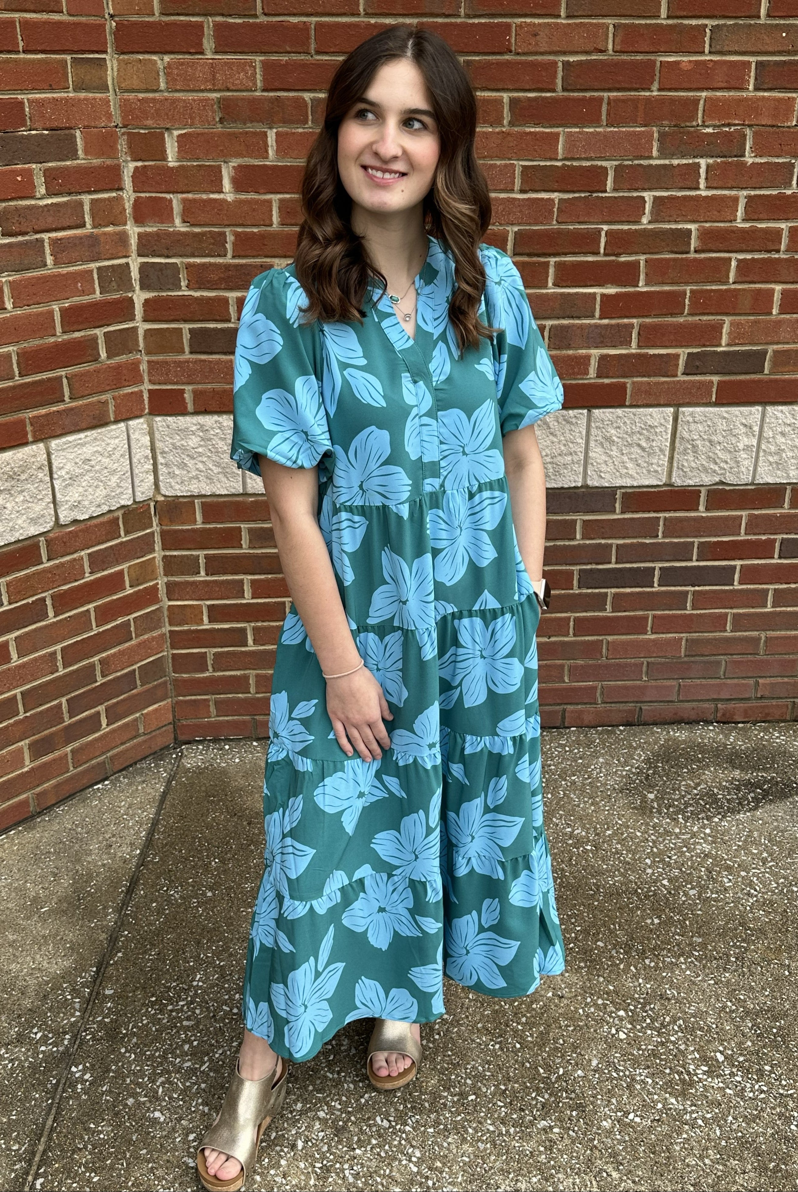 Woman wearing a teal floral dress standing against a brick wall.