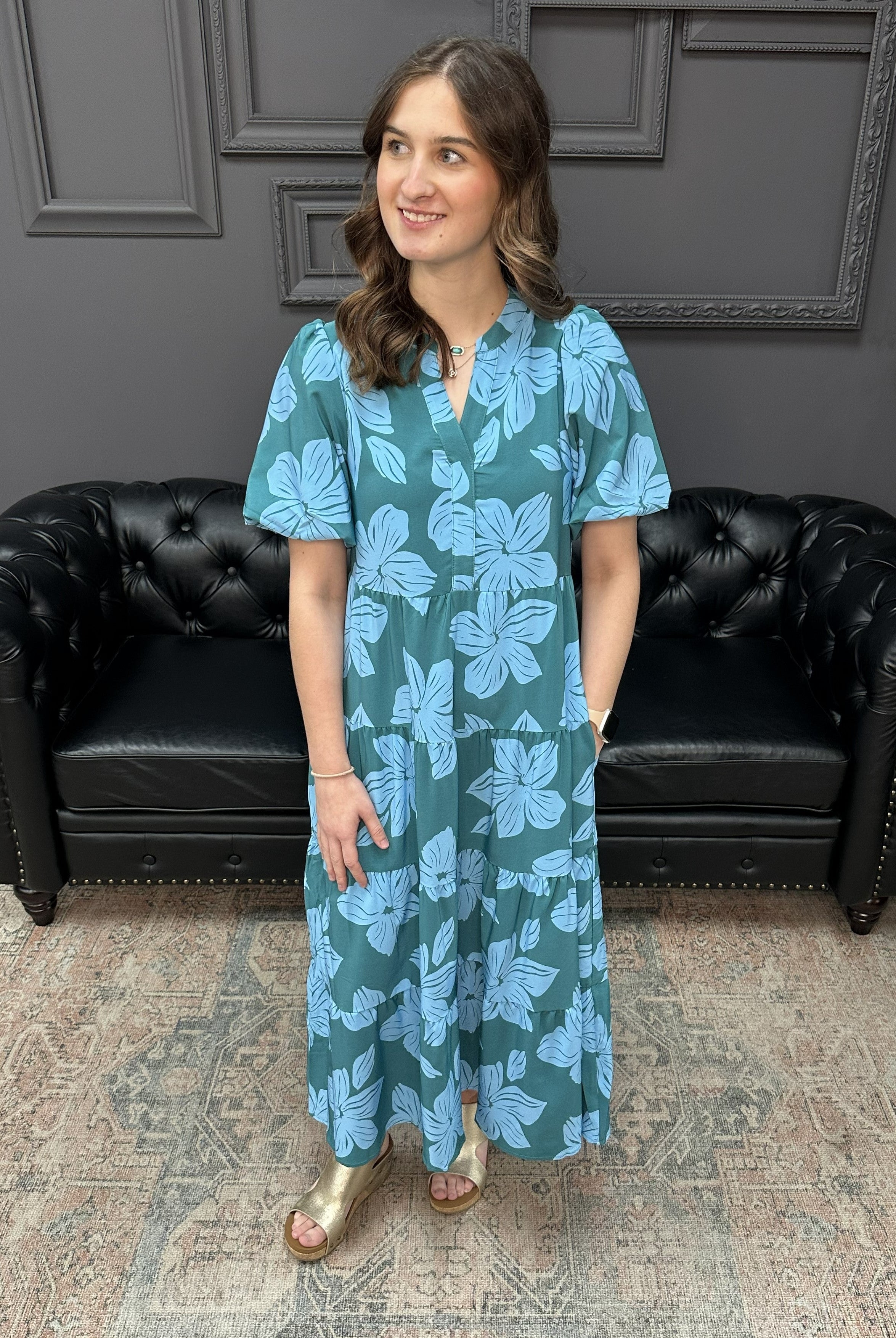 Woman wearing a blue floral dress standing in a room with a black leather couch and gray walls.