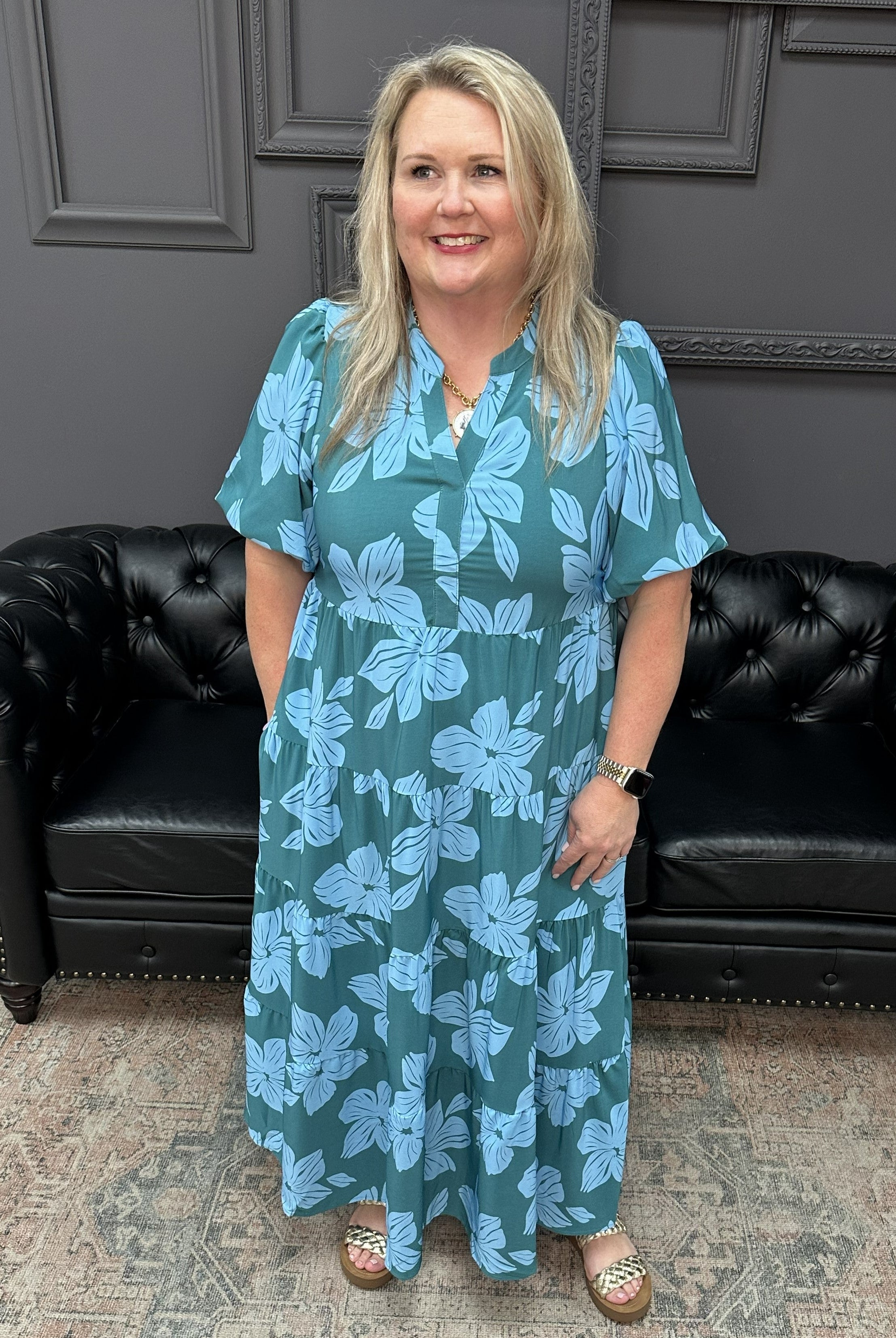 Woman in a teal floral dress standing in a room with black leather couches and a patterned rug.