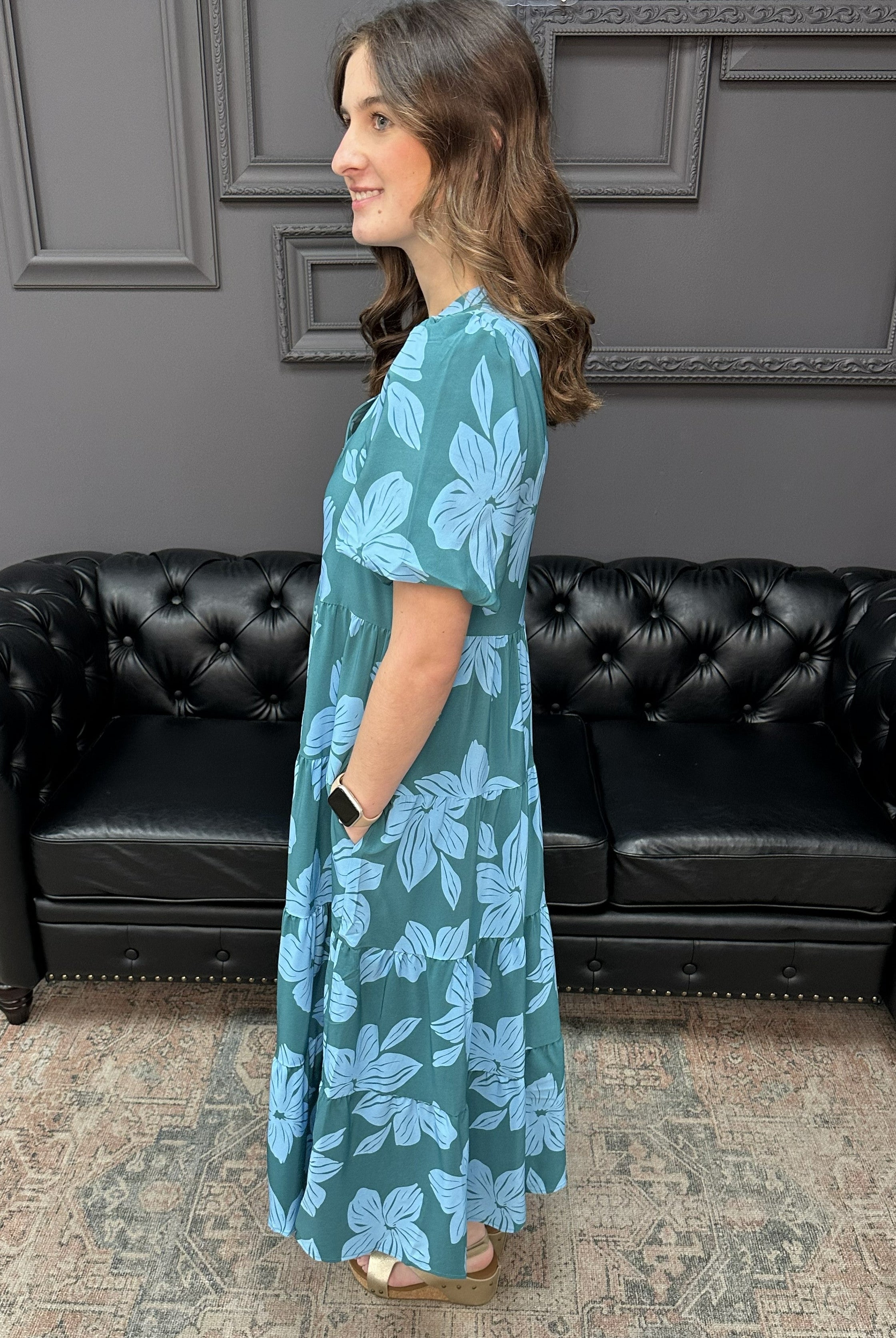 Woman wearing a blue floral dress standing in a room with a black leather couch and gray walls.