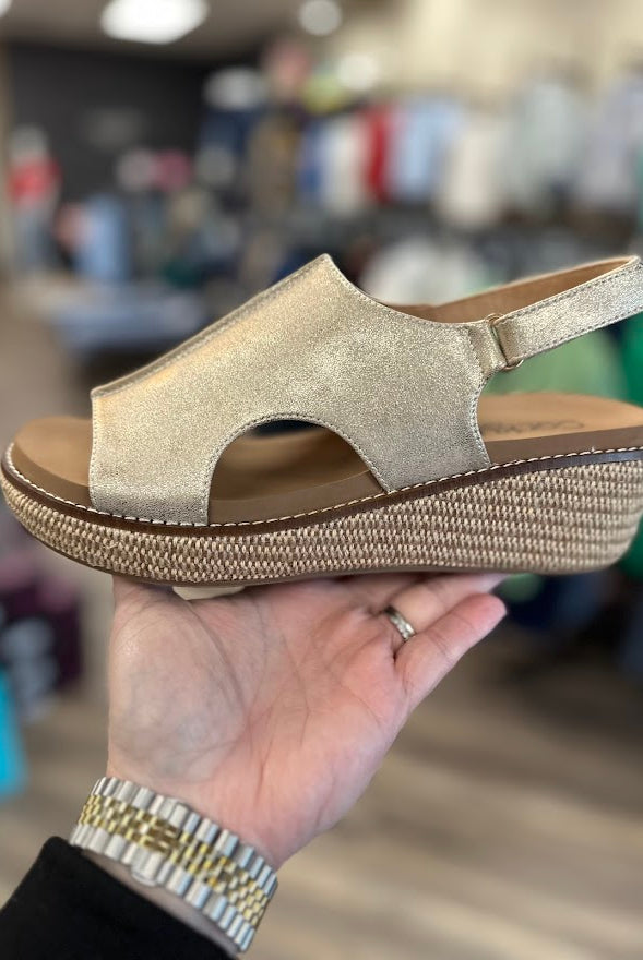 Gold wedge sandal held in a hand with a blurred store background