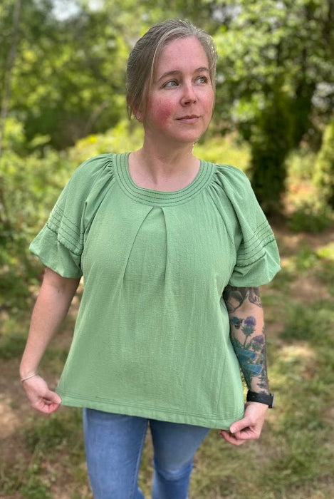 Woman wearing a green blouse and blue jeans standing outdoors with trees in the background
