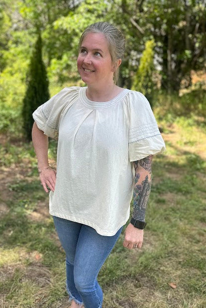 Woman standing outdoors in a natural setting wearing a light-colored blouse and blue jeans.