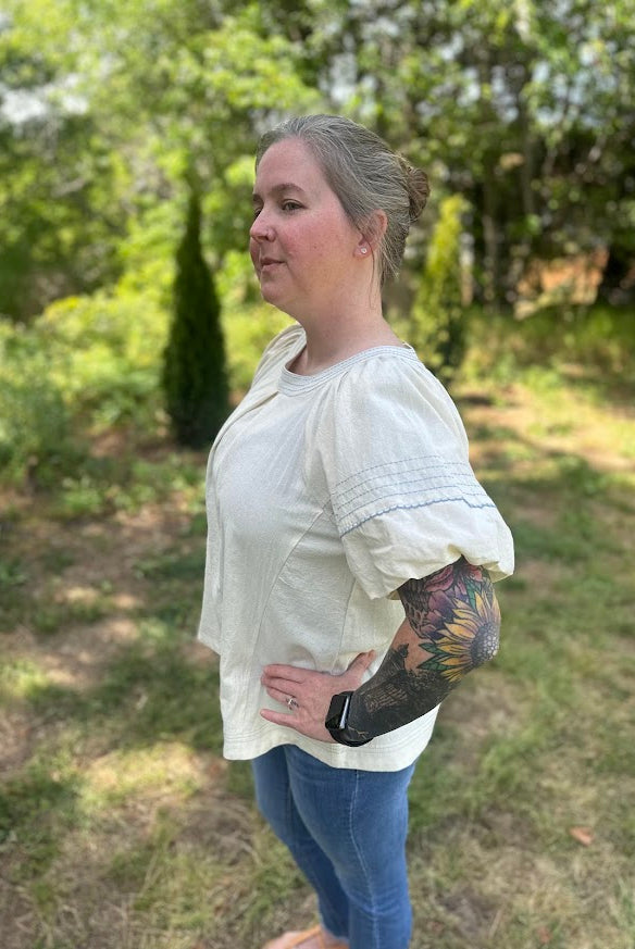 Woman wearing a white blouse and blue jeans standing outdoors with greenery in the background