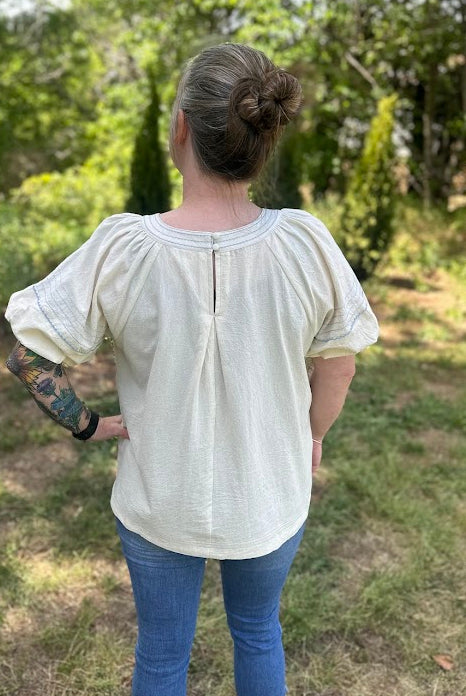 Person wearing a white blouse and blue jeans standing in a grassy area with trees.