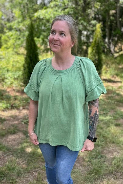 Person wearing a green shirt and blue jeans standing in a grassy outdoor area.