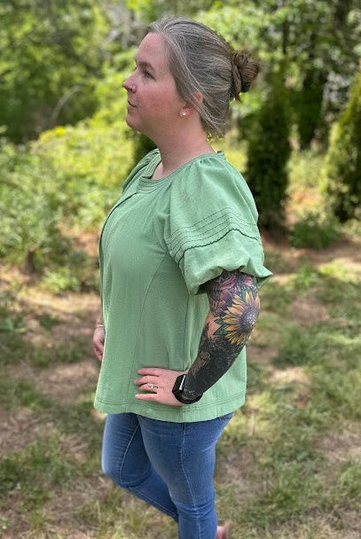 Woman wearing a green blouse and blue jeans standing in a grassy area with trees.
