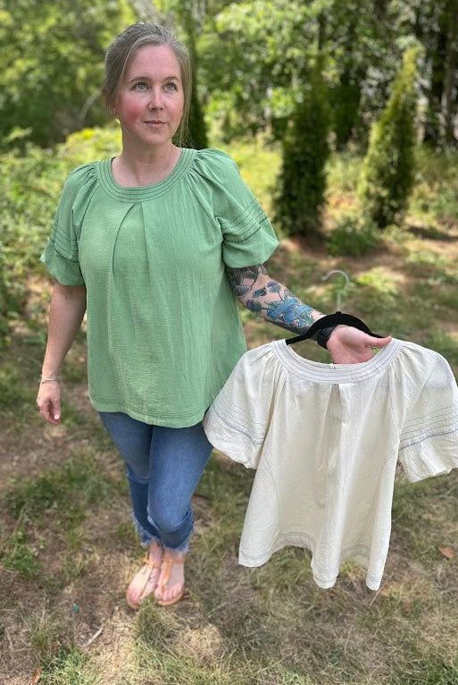 Woman holding a white blouse outdoors with greenery in the background