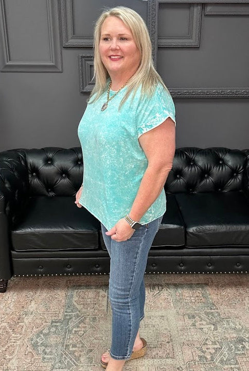 Woman in a teal tie-dye shirt and jeans standing in front of a black leather couch.