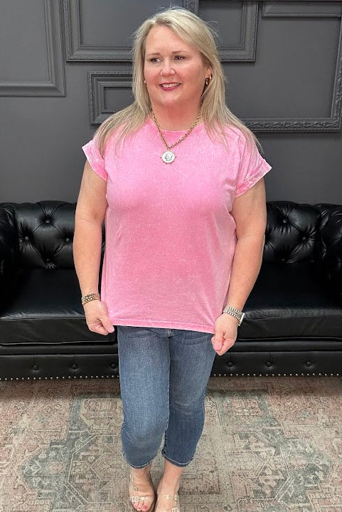 Woman in a pink shirt standing in front of a black leather couch with a gray wall and neon sign in the background.