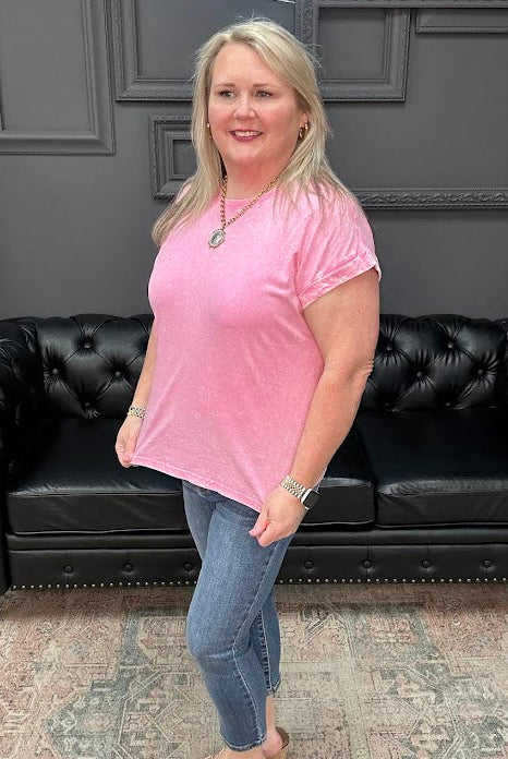 Woman in a pink shirt standing in front of a black leather couch with a gray wall and neon sign in the background.
