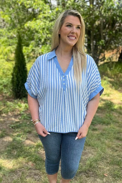Woman wearing a blue and white striped shirt and jeans standing in a grassy area with trees.