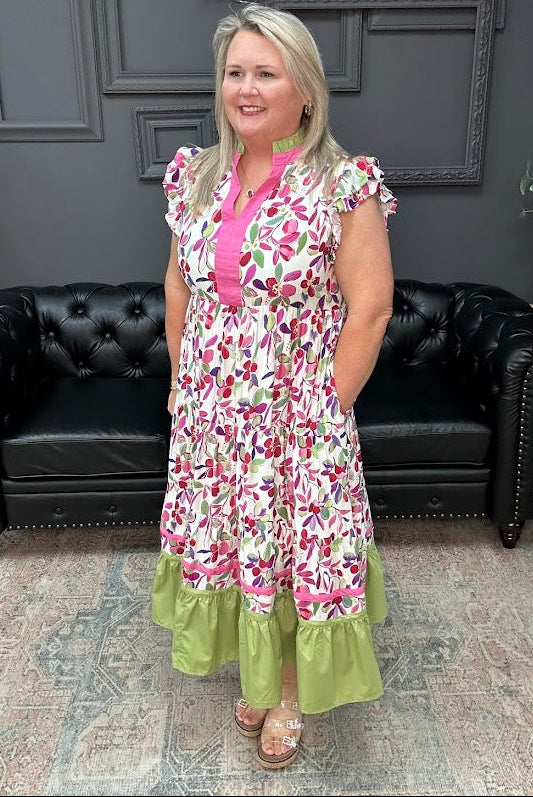 Woman wearing a floral dress with a pink top and green bottom in a room with black leather couches and gray walls.