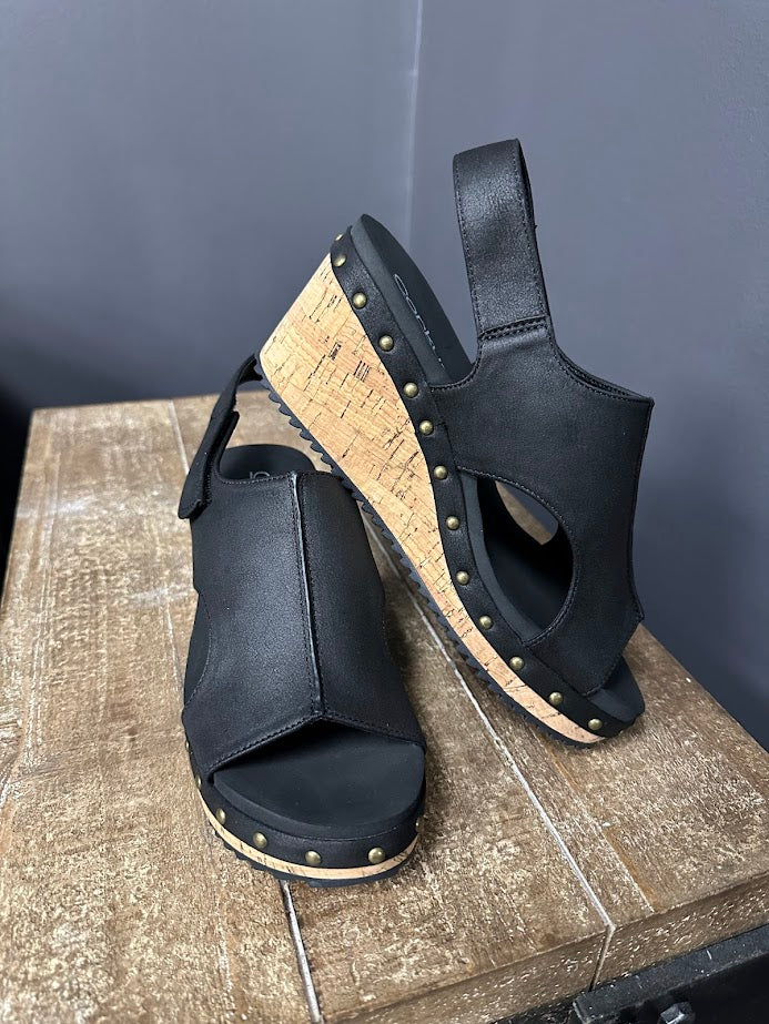 Black open-toe sandals with cork insole on a wooden surface against a gray wall.