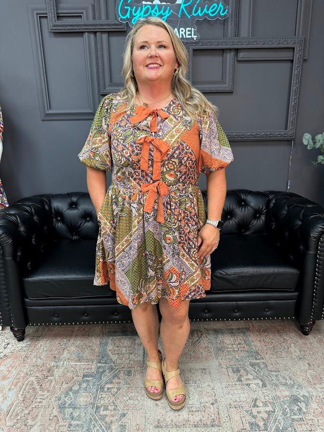 Woman in a patterned dress standing in front of a black leather couch with a gray wall and decorative elements in the background.