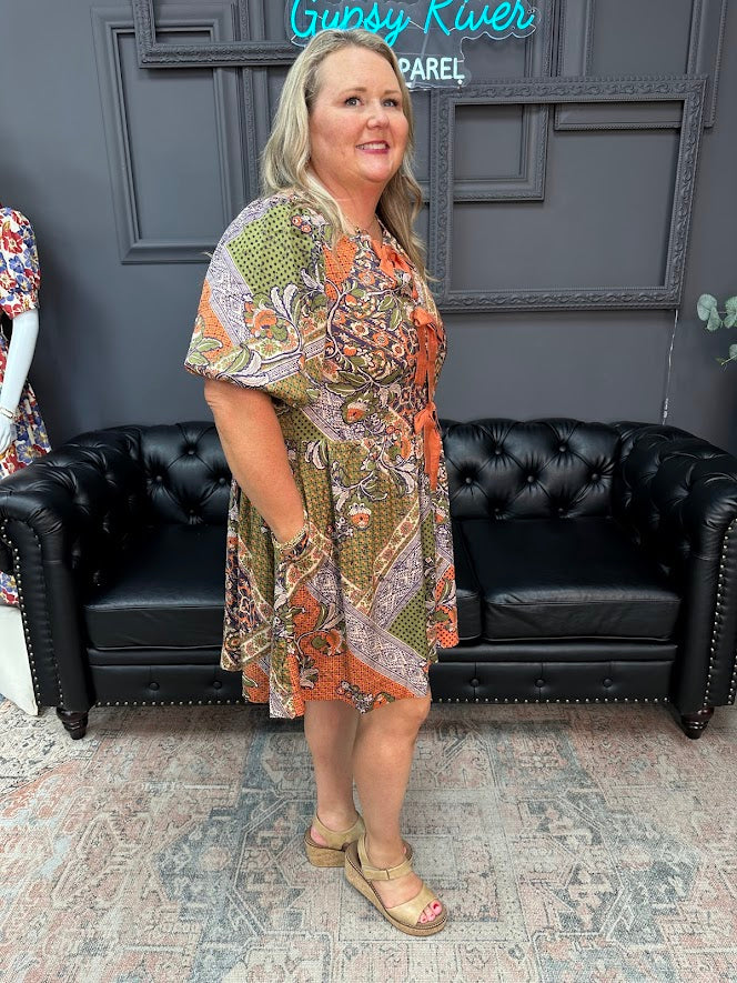 Woman in a patterned dress standing in front of a black leather couch with a dark gray wall and 'Gunsy River Apparel' sign in the background.