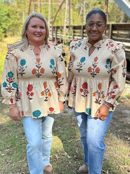 Two women wearing matching embroidered tops standing in a grassy area with a bridge in the background.
