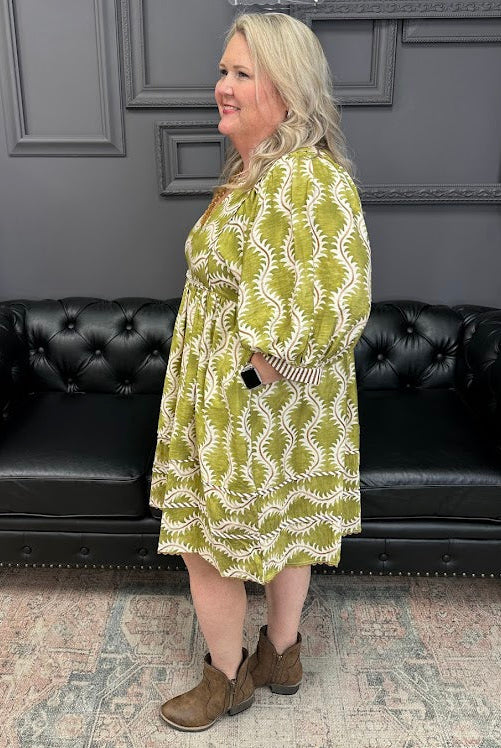 Woman wearing a green and white patterned dress standing in front of a black leather couch.