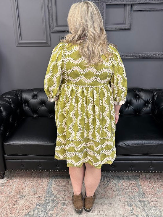 Woman wearing a green patterned dress standing in front of a black leather couch with 'Gypsy River' branding.