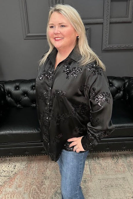 Woman wearing a black embellished jacket standing in a room with a black couch and framed pictures on the wall.