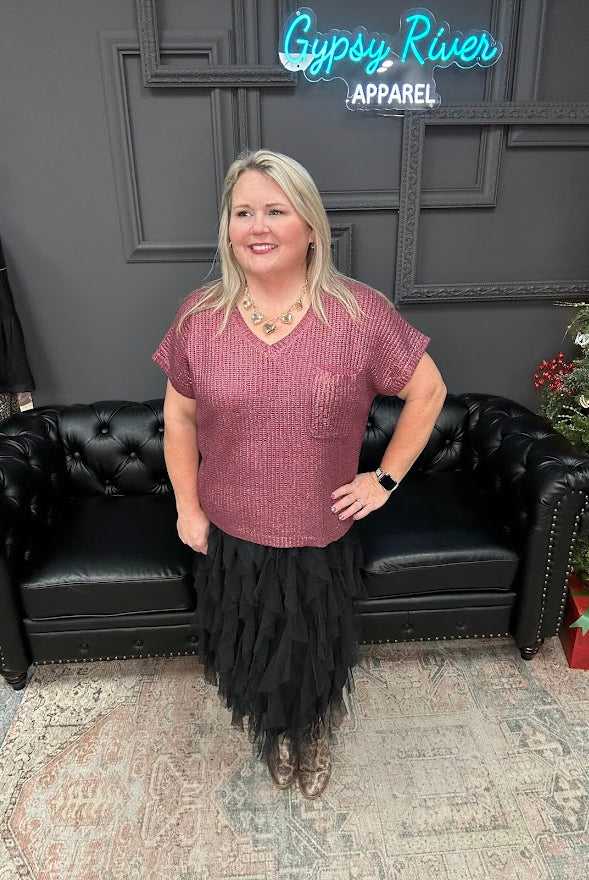 Woman posing in a room with a black couch and Christmas tree, wearing a maroon top and black skirt.
