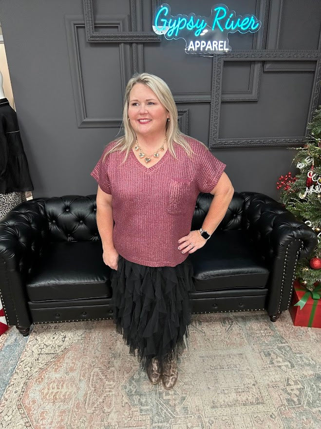 Woman posing in a room with a black couch and Christmas tree, wearing a maroon top and black skirt.