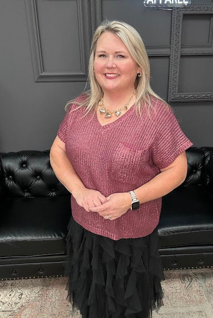 Woman in a pink top and black skirt standing in front of a black leather couch with a Christmas tree and 'Gypsy River Apparel' sign in the background.