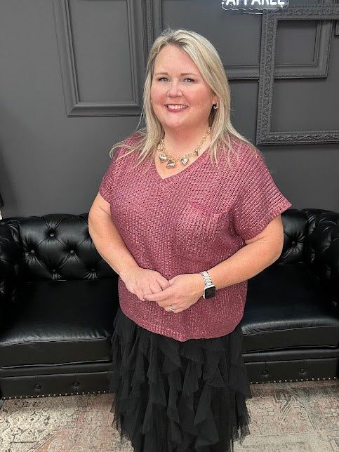 Woman in a pink top and black skirt standing in front of a black leather couch with a Christmas tree and 'Gypsy River Apparel' sign in the background.