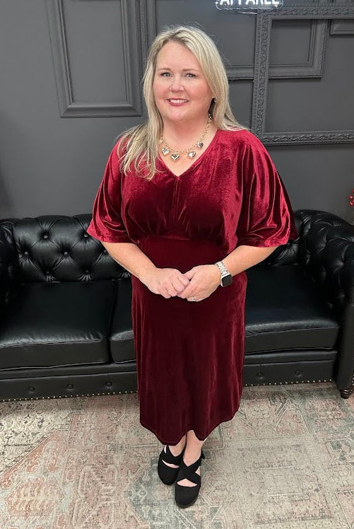 Woman in a burgundy dress standing in front of a black leather couch with 'Gypsy River Apparel' sign.