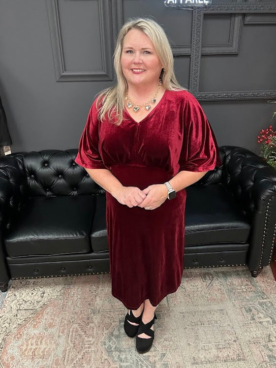 Woman in a burgundy dress standing in front of a black leather couch with 'Gypsy River Apparel' sign.