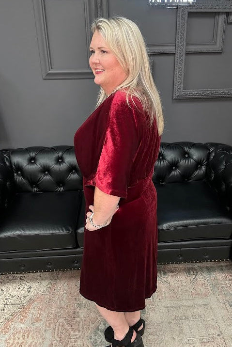 Woman in a burgundy dress standing in front of a black leather couch with 'Gypsy River Apparel' branding.