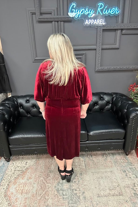 Person wearing a red velvet dress standing in front of a black leather couch with 'Gypsy River Apparel' branding.