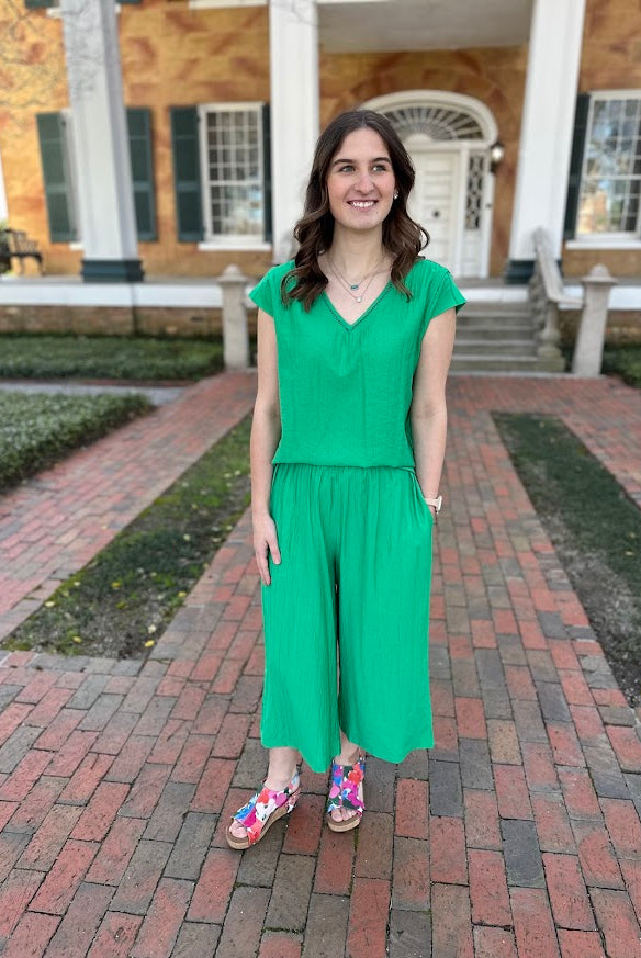 Woman in a green jumpsuit standing on a brick path in front of a building.
