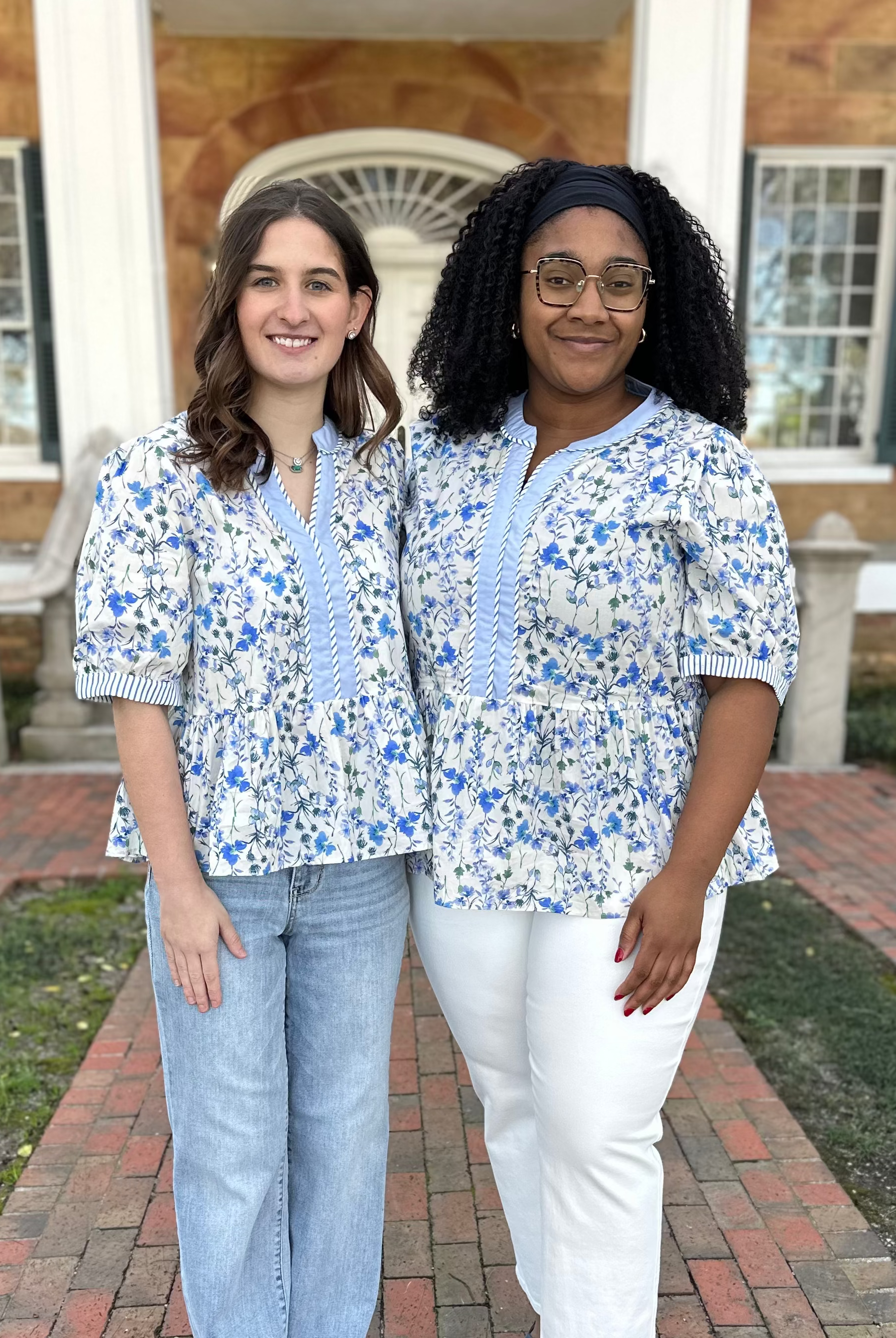 Two women in matching floral shirts standing on a brick path in front of a building.