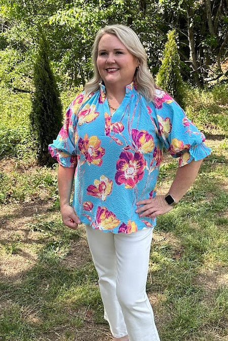 Woman wearing a colorful floral blouse and white pants standing in a grassy area with trees.