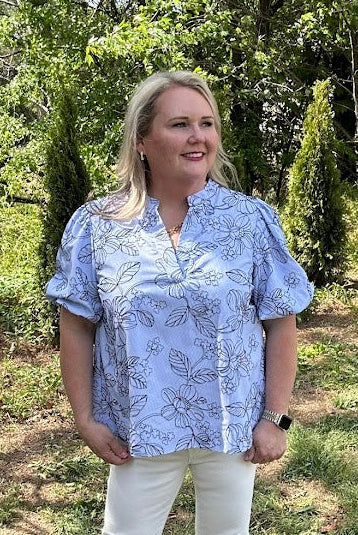 Woman standing outdoors in a blue floral blouse and white pants, surrounded by greenery.