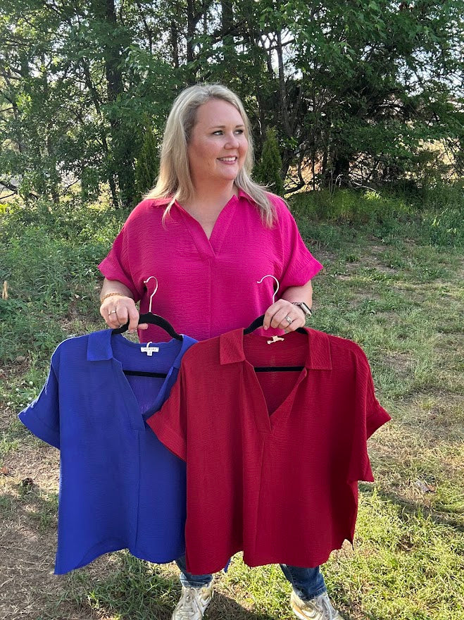 Woman holding two shirts, one blue and one red, outdoors with trees in the background.