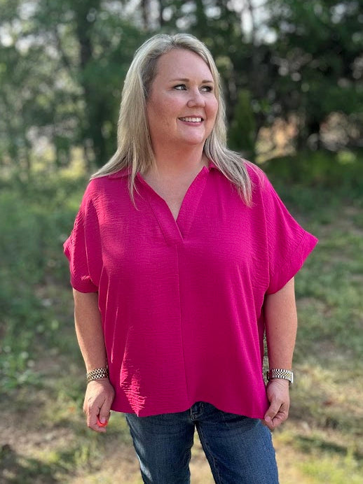 Woman wearing a bright pink top and blue jeans standing outdoors with trees in the background