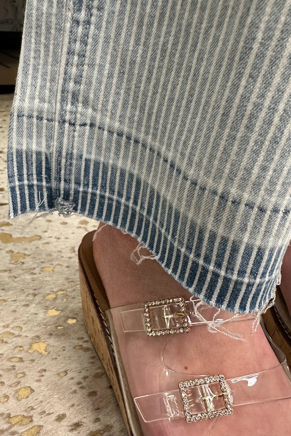 Close-up of striped jeans and clear sandals with rhinestone buckles on a textured floor.