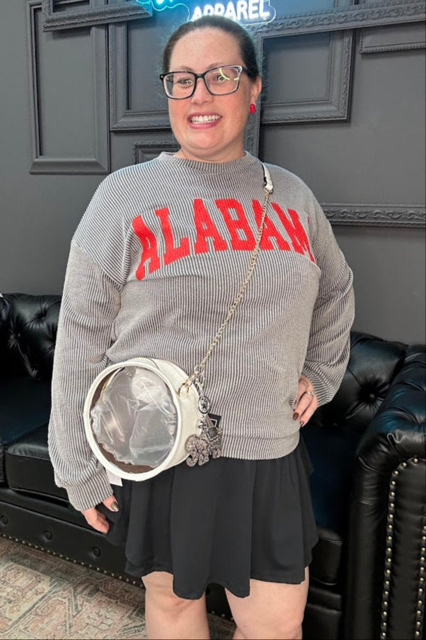 Woman wearing a gray sweater with red text, holding a round handbag, standing in a room with dark walls and a black leather couch.