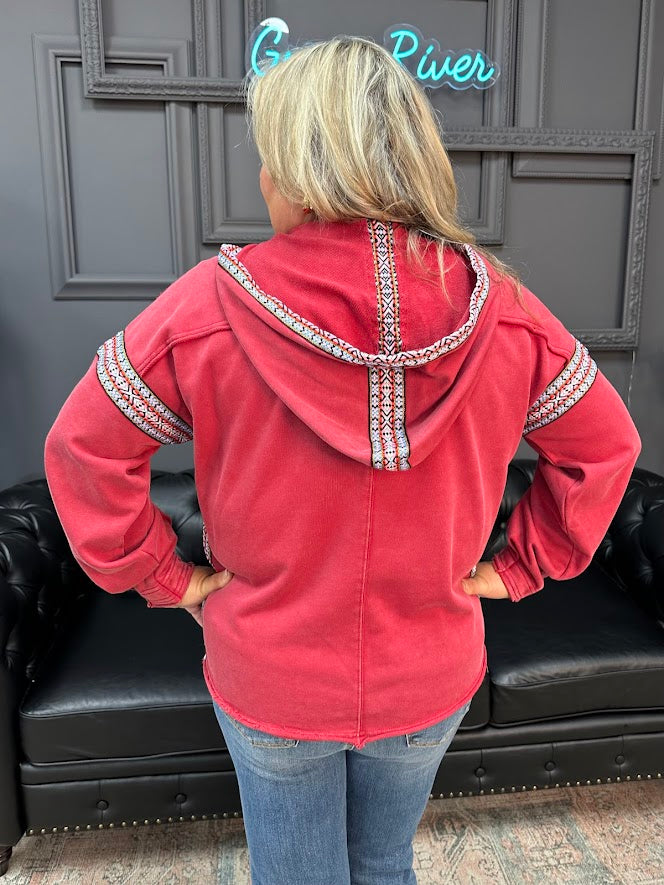 Person wearing a red hoodie with decorative trim, standing in front of a black leather couch and gray wall.