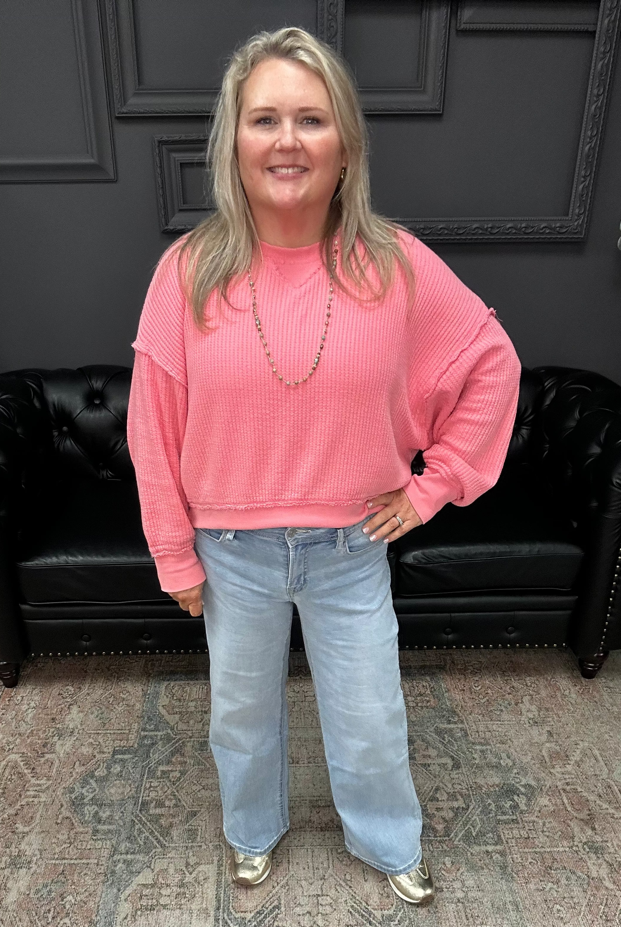 Woman wearing a pink sweater and blue jeans standing in a room with dark walls and a black leather couch.