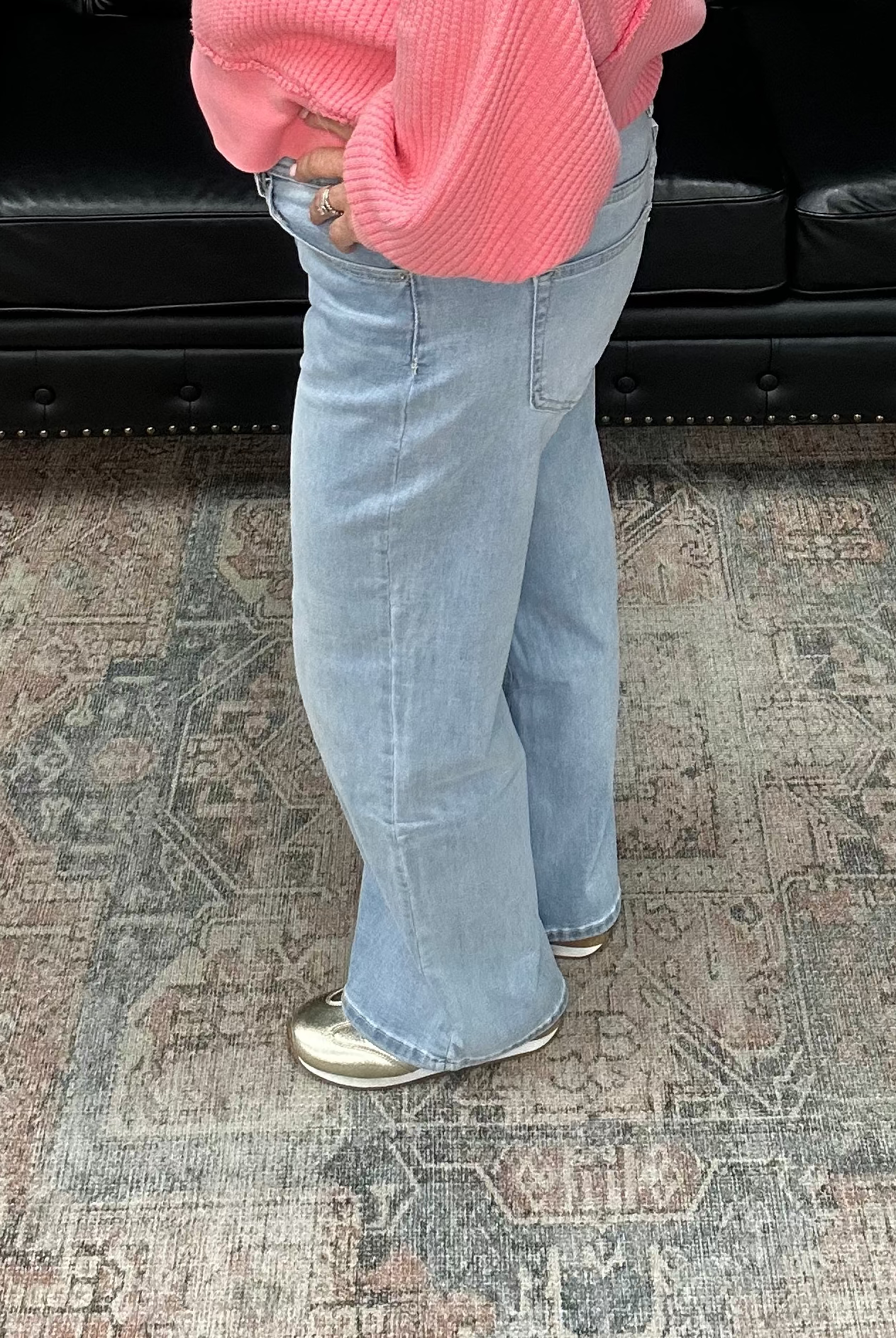 Woman in a pink sweater and blue jeans standing in a room with a black leather sofa and decorative items.