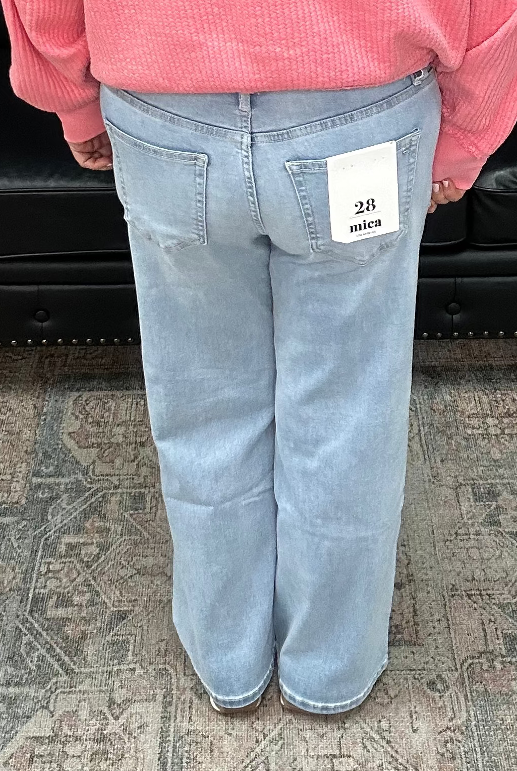 Person wearing a pink sweater and light blue jeans standing in a room with a black leather sofa and patterned rug.