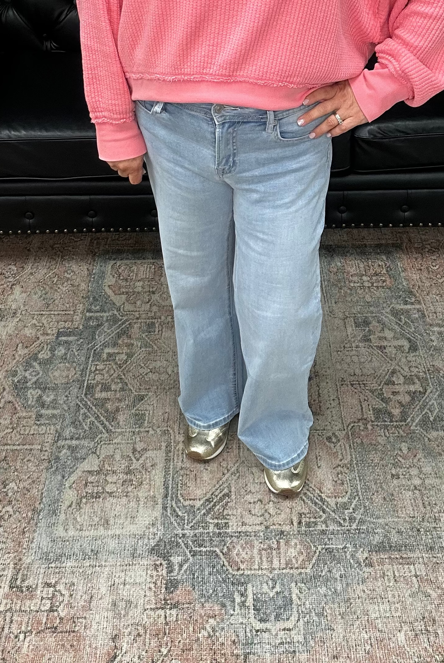 Woman wearing a pink sweater and blue jeans standing in a room with a black leather couch and floral rug.
