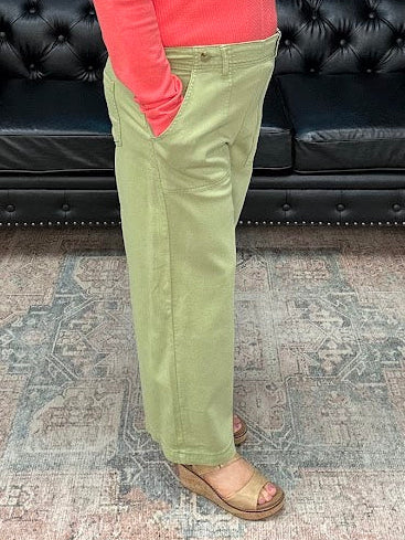 Woman in a pink top and green pants standing in a room with a black leather couch and floral decorations.