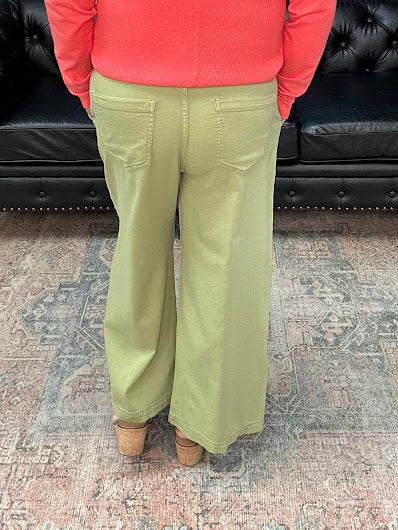 Person wearing a coral sweater and green pants standing in a room with a black leather couch and floral decorations.